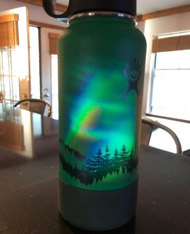 Zelená fľaša s lesným motívom a dúhovým odrazom svetla na kuchynskom stole.