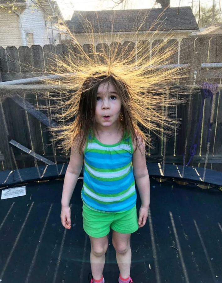 Dievčatko so statickou elektrinou vo vlasoch na trampolíne, oblečené v pruhovanom tričku a zelených šortkách.