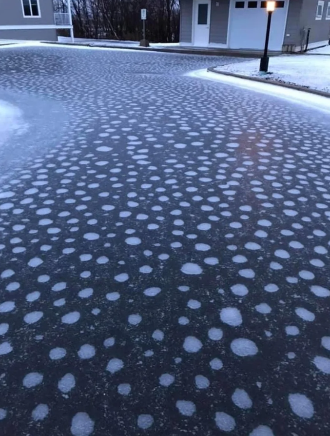 Zasnežená cesta s bielymi škvrnami na asfalte pred domom, zimná krajina.