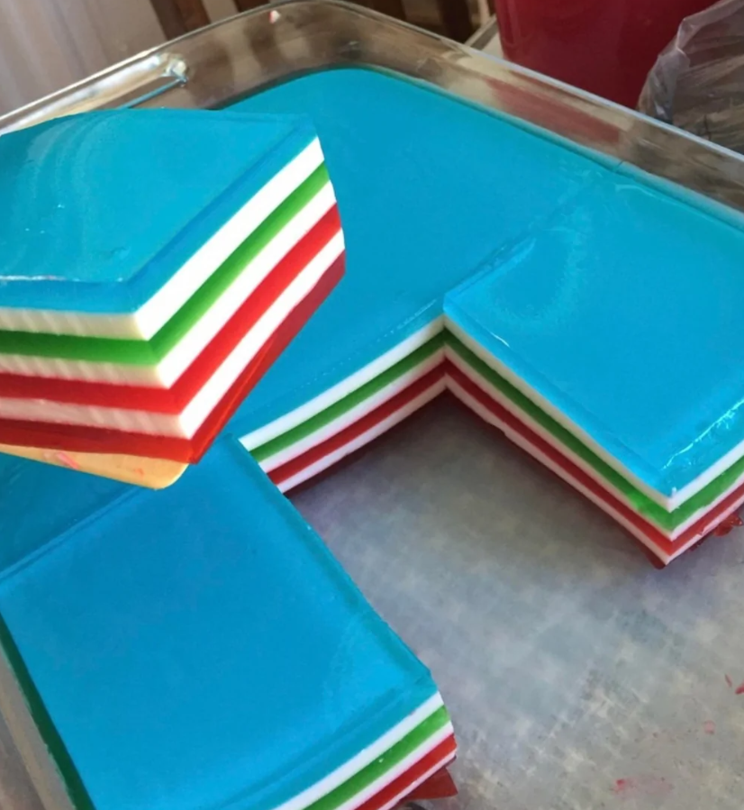 Viacfarebná želé torta s vrstvami modrej, bielej, zelenej a červenej na podnose, so štvorcovým výrezom.