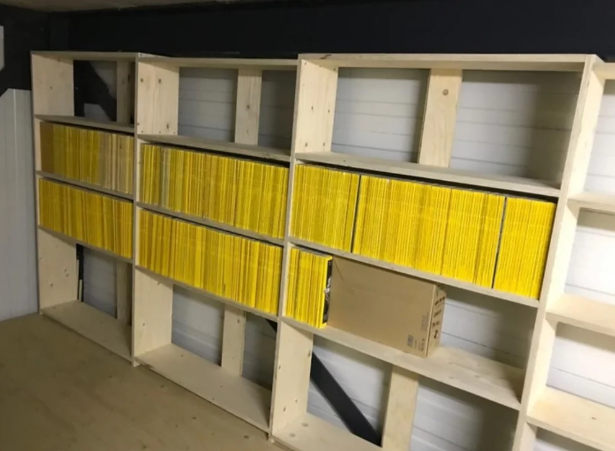 Polička plná žltých časopisov so škatuľou v spodnom rade, v drevenom regáli na bielom pozadí.