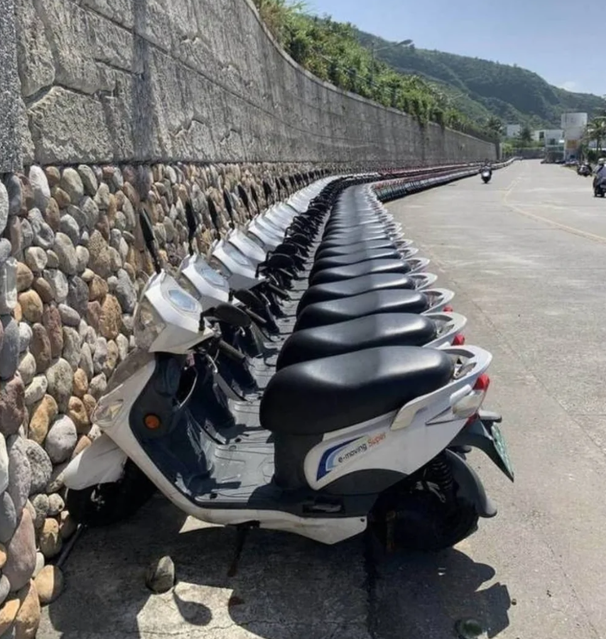 Riadok skútrov zaparkovaných vedľa kamenného múru na slnečnej ulici s horami na pozadí.