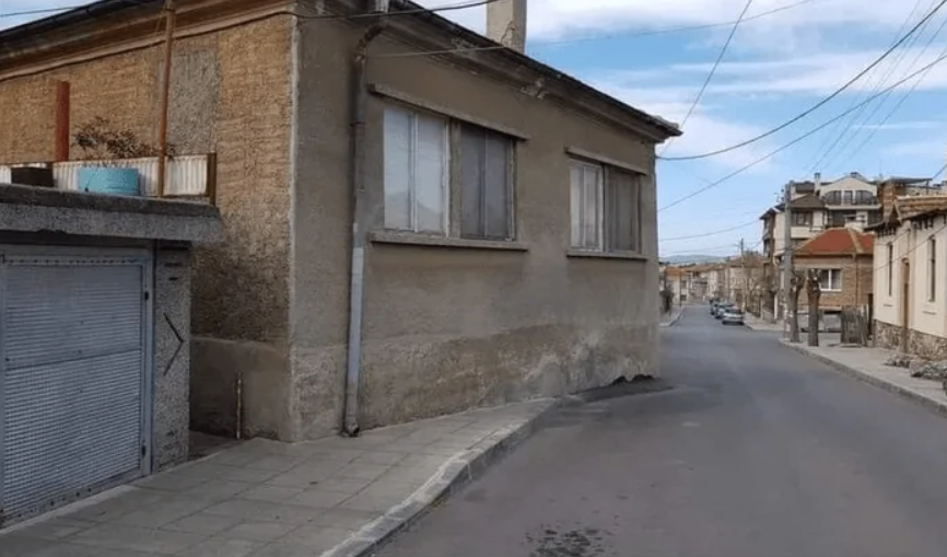 Ulica s domom zasahujúcim do vozovky, nebom posiatym oblakmi a okolitými budovami na pokojnej mestskej scéne.