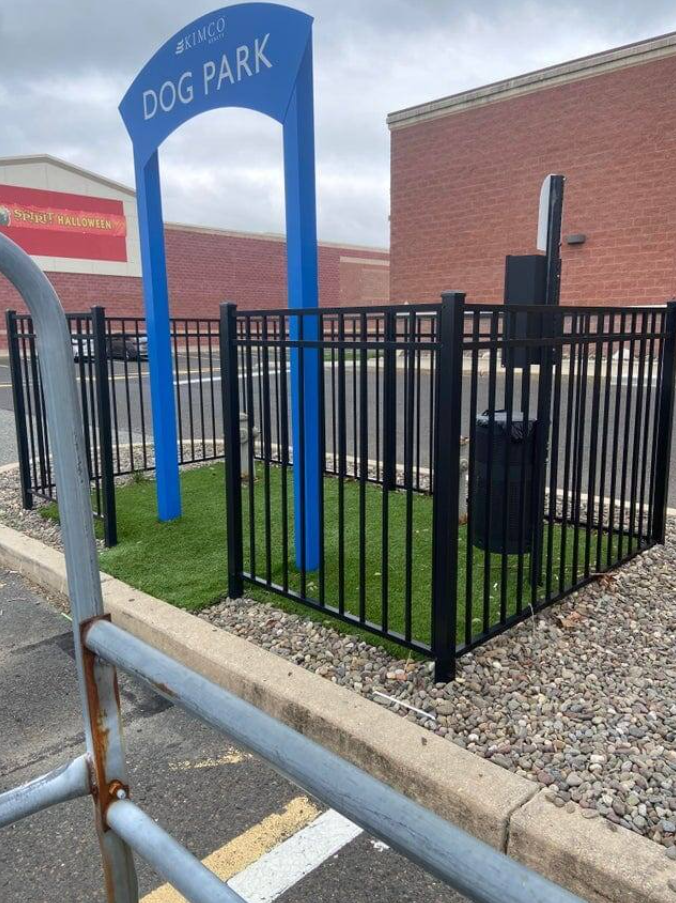 Malý oplotený priestor označený nápisom Dog Park so zeleňou a odpadkovým košom na parkovisku.