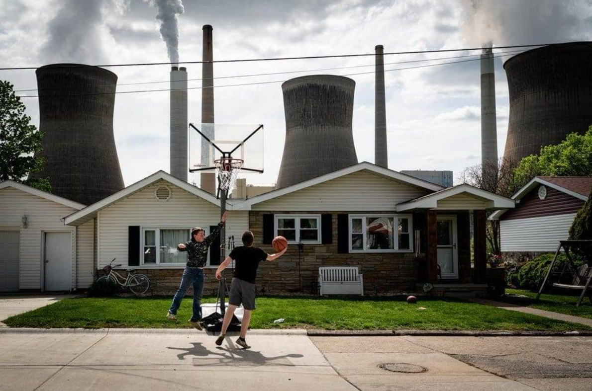 Deti hrajú basketbal pred domom, v pozadí sú chladiace veže elektrárne, symbol kontrastu medzi prírodou a priemyslom.