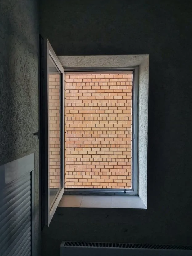 Okno v tmavej miestnosti s výhľadom na tehlovú stenu vonkajšej budovy.