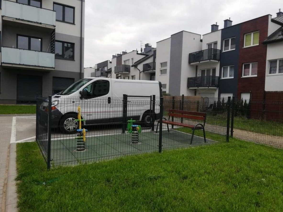 Dodávka parkuje vedľa oploteného detského ihriska v bytovej štvrti s modernými budovami.