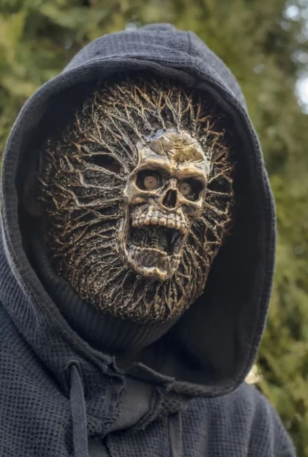 Osoba v tmavej mikine s kapucňou nosí výraznú masku s lebkovým motívom v prírodnom prostredí.