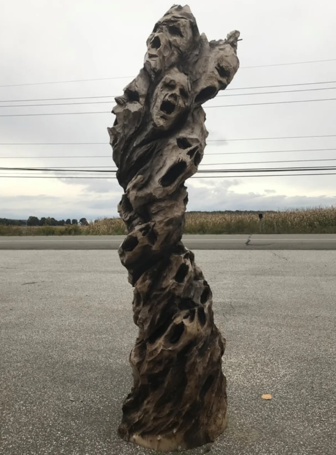 Drevená socha s vyrezávanými tvárami na parkovisku pod oblačnou oblohou.