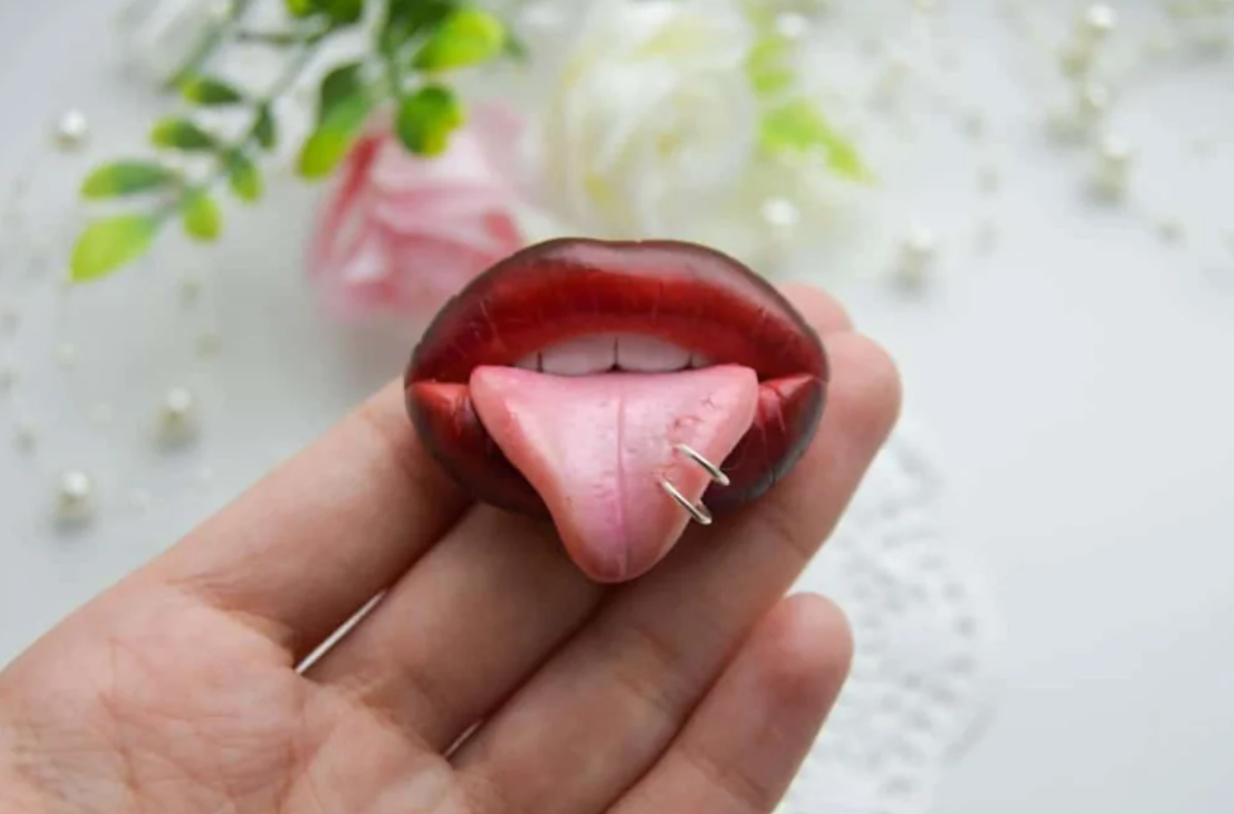 Ružové pery s vypľazeným jazykom, ozdobeným prsteňom, držiace na ruke, na pozadí kvety.