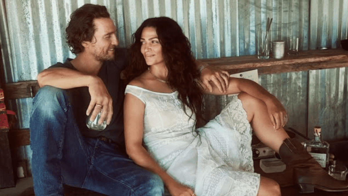 Pár sedí na lavičke, muž drží pohár, oblečený v džínsach, žena v bielej letnej šatách s kovbojskými čižmami.