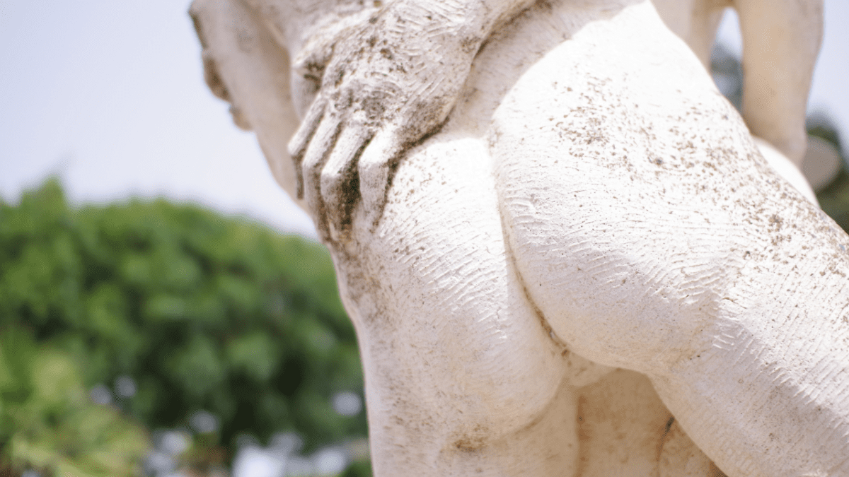 Socha v parku s detailmi rúk a kameňa, zaostrená na textúru na pozadí zeleného lístia.