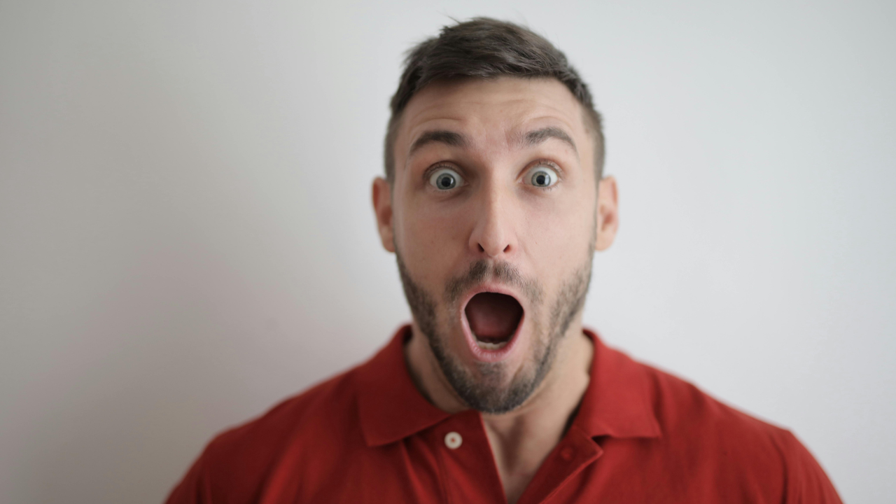 Muž v červenom tričku s prekvapeným výrazom tváre a otvorenými ústami na svetlom pozadí.