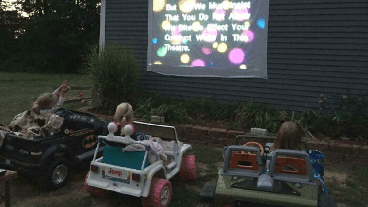 Deti sledujú film na projektore vonku, sediac v malých autíčkach na dvore.