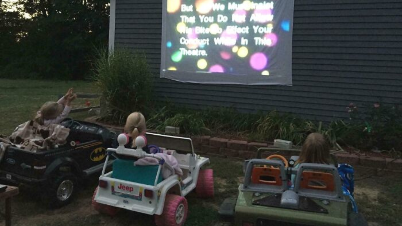 Deti sledujú film na projektore vonku, sediac v malých autíčkach na dvore.