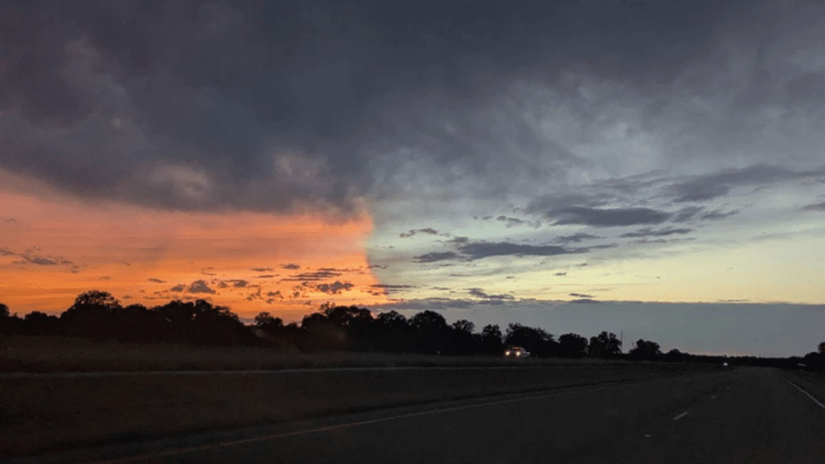 Cesta počas západu slnka, obloha s farebným kontrastom oranžovej a fialovej so siluetami stromov na horizonte.