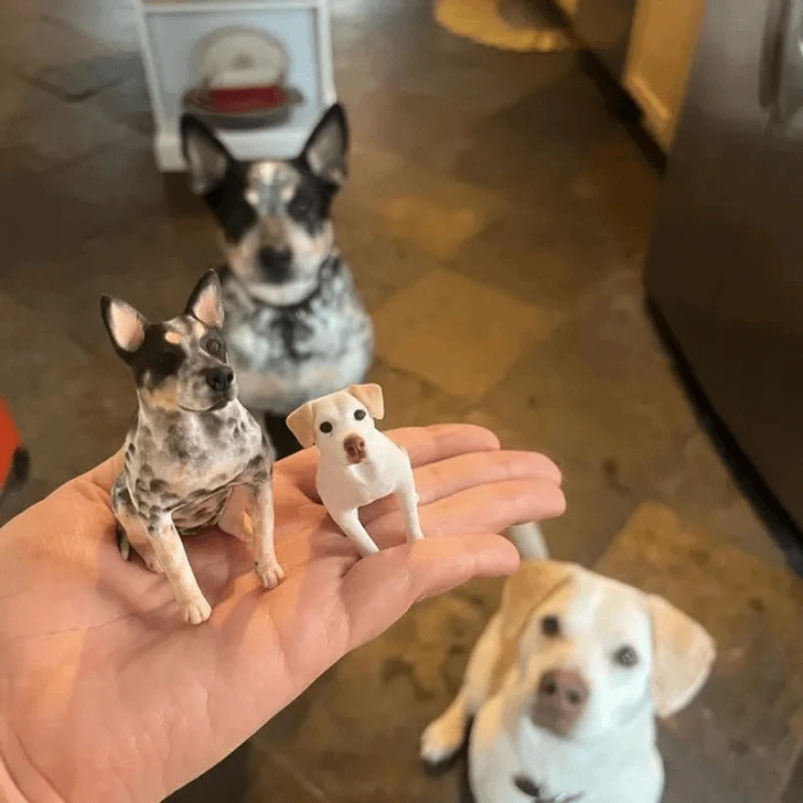 Miniatúrne modely psov na dlani, v pozadí skutoční psi na kuchynskej podlahe.