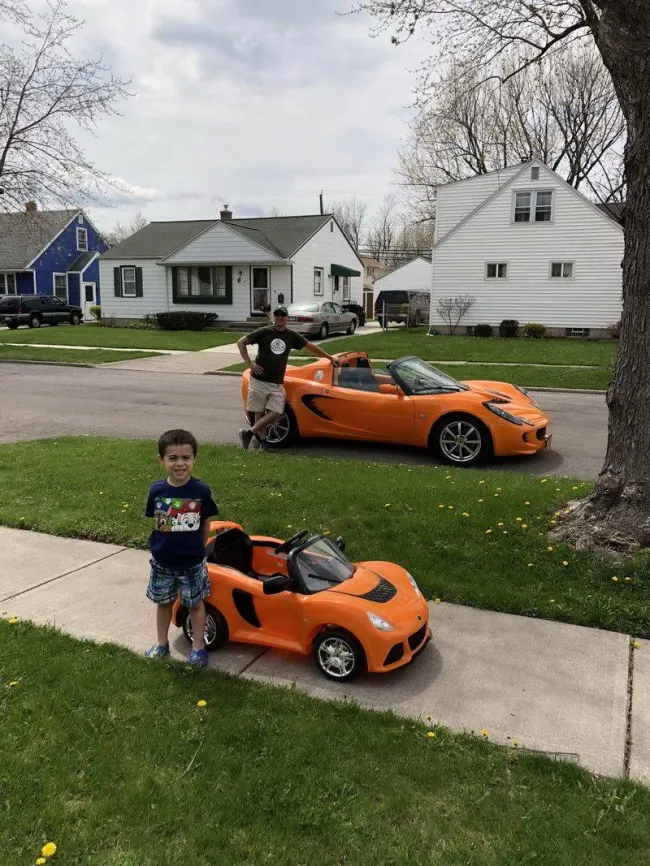 Otec a syn s oranžovými autíčkami, jedno veľké a jedno malé, pred domom na zelenej tráve.