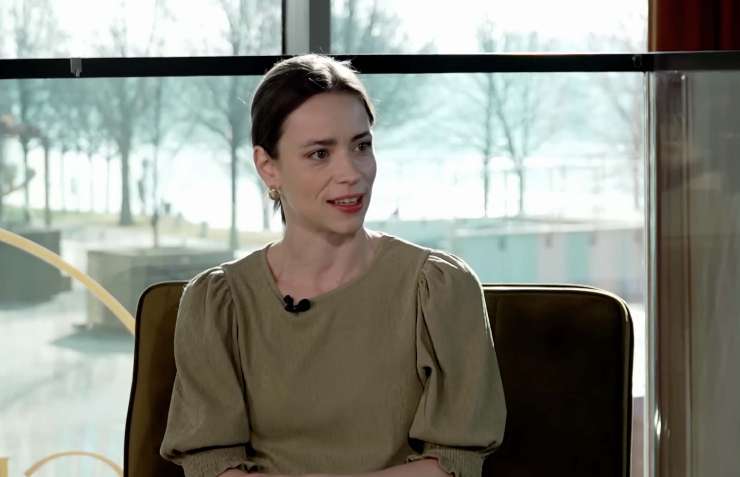 Neznáma žena v béžovom oblečení sedí vo vnútri, usmieva sa, v pozadí sú okná s výhľadom na vonkajšiu scenériu.