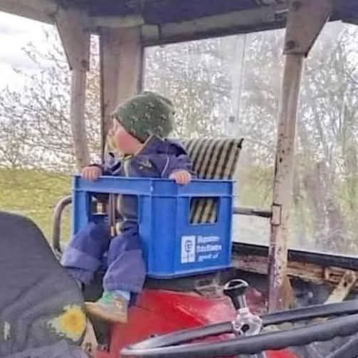 Dieťa sedí v plastovej debničke v traktore na farme, oblečené v teplej bunde a čiapke.