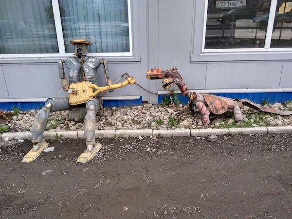 Robot hrajúci na gitare vedľa sochy draka pred sivou budovou.