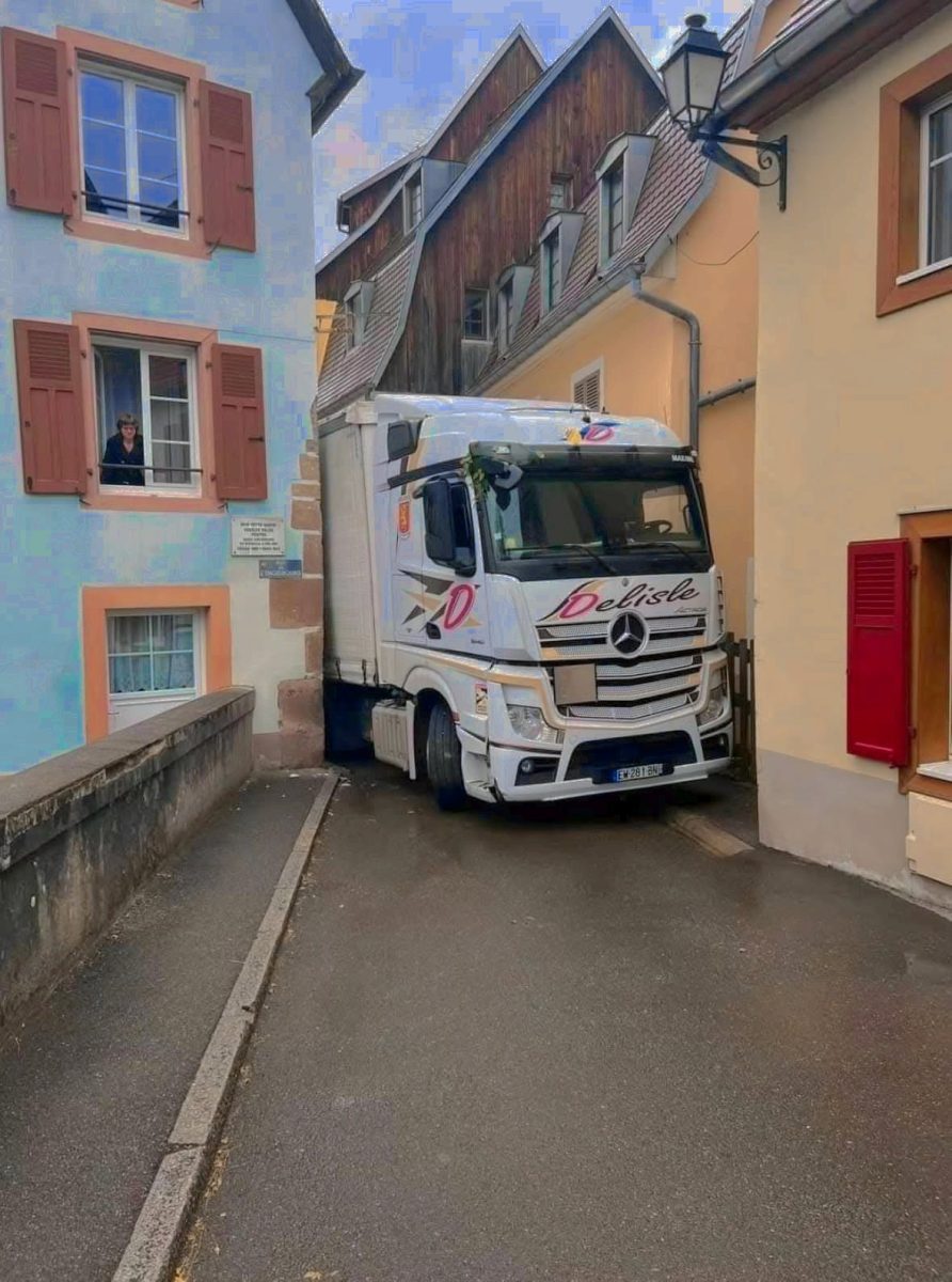 Kamión zaseknutý v úzkej uličke medzi domami, okoloidúci sleduje situáciu z okna.