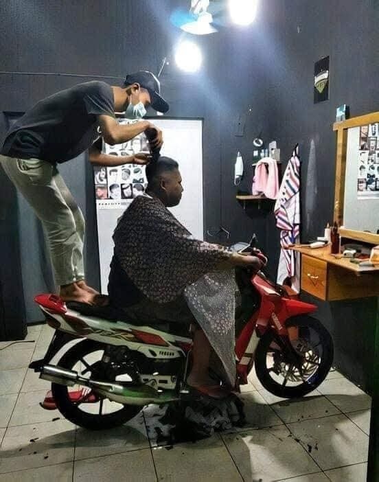 Muž dostáva účes na motorke v interiéri, kaderník stojí na sedadle. Kombinácia kreativity a pohodlia.