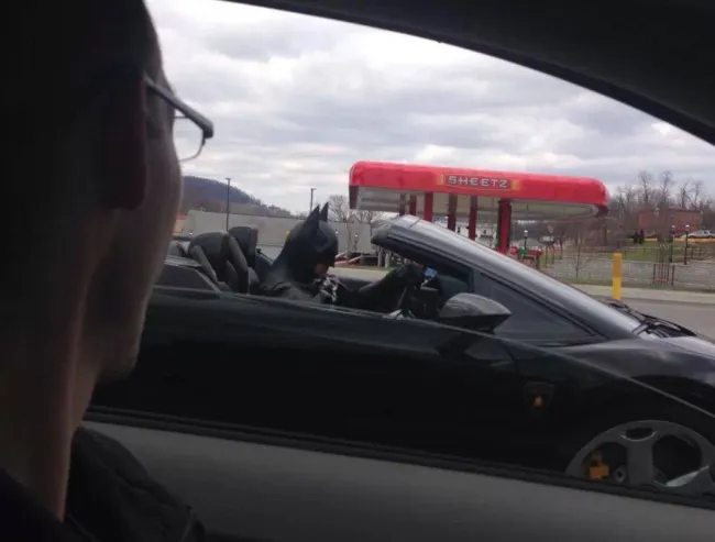 Muž v Batmanovom kostýme v športovom aute naštartovaný pri čerpacej stanici, pohľad z vedľajšieho auta.