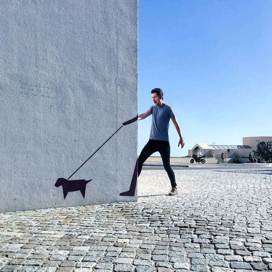 Muž interaguje s nástennou maľbou psa na chodníku, vytvárajúcej dojem venčenia.
