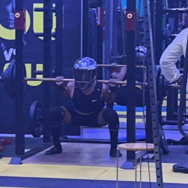 Osoba v prilbe robí drep s činkou v posilňovni, obklopená tréningovým zariadením.