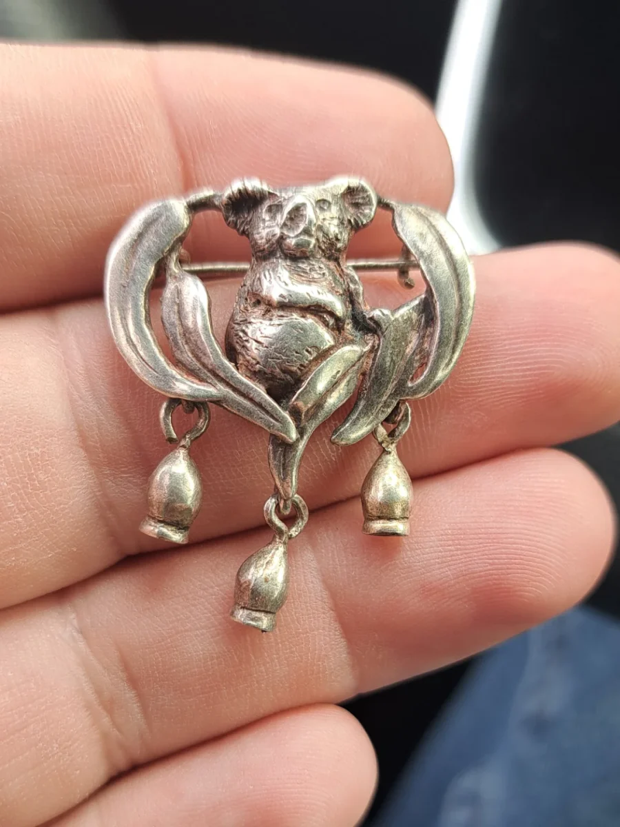 Strieborná brošňa s motívom koaly a eukalyptových listov, držiaca na prstoch ruky.