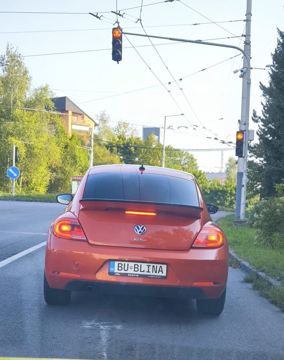 Oranžový Volkswagen Beetle s personalizovanou EČV na červenej na semafore, letný deň.
