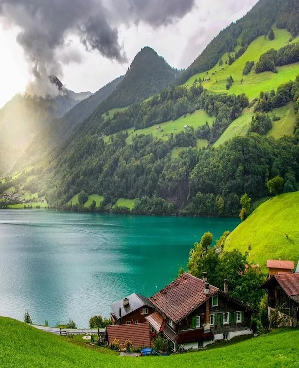 Horské jazero obklopené zelenými svahmi a chatkami pod dramatickými mrakmi. Tichá alpská scenéria na vidieku.