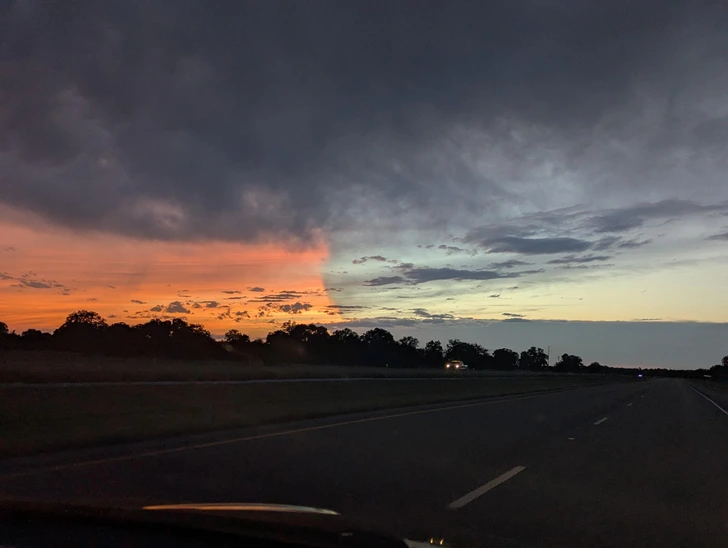 Cesta za súmraku s dramatickým prechodom z oranžovej do tmavej oblohy, siluety stromov na horizonte.