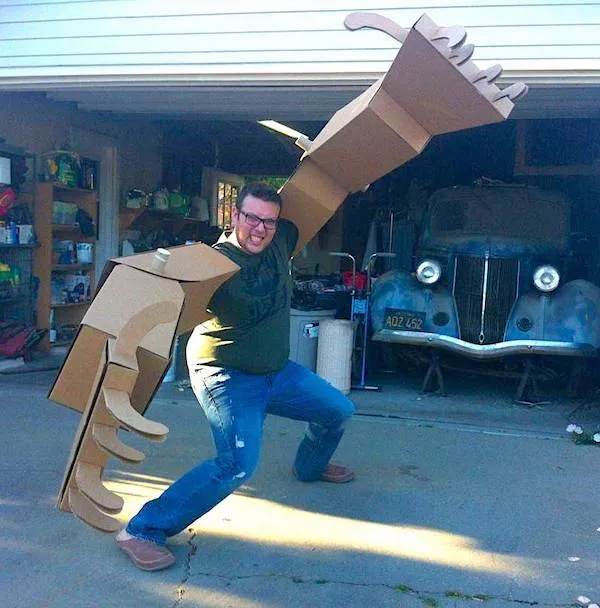 Osoba pózuje s obrovskými kartónovými ramenami pred garážou so starým autom. Kreatívny moment so zábavným nádychom.