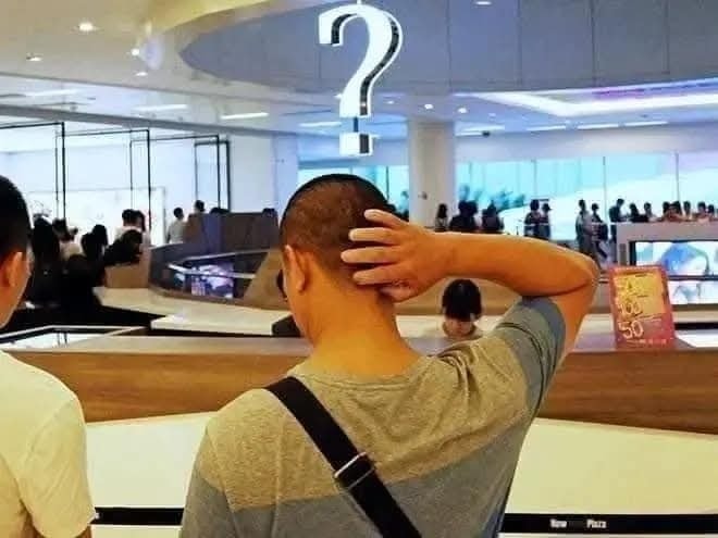 Muž v nákupnom centre stojí pod veľkým otáznikom, držiac sa za hlavu, čo vyvoláva dojem zmätku alebo premýšľania.