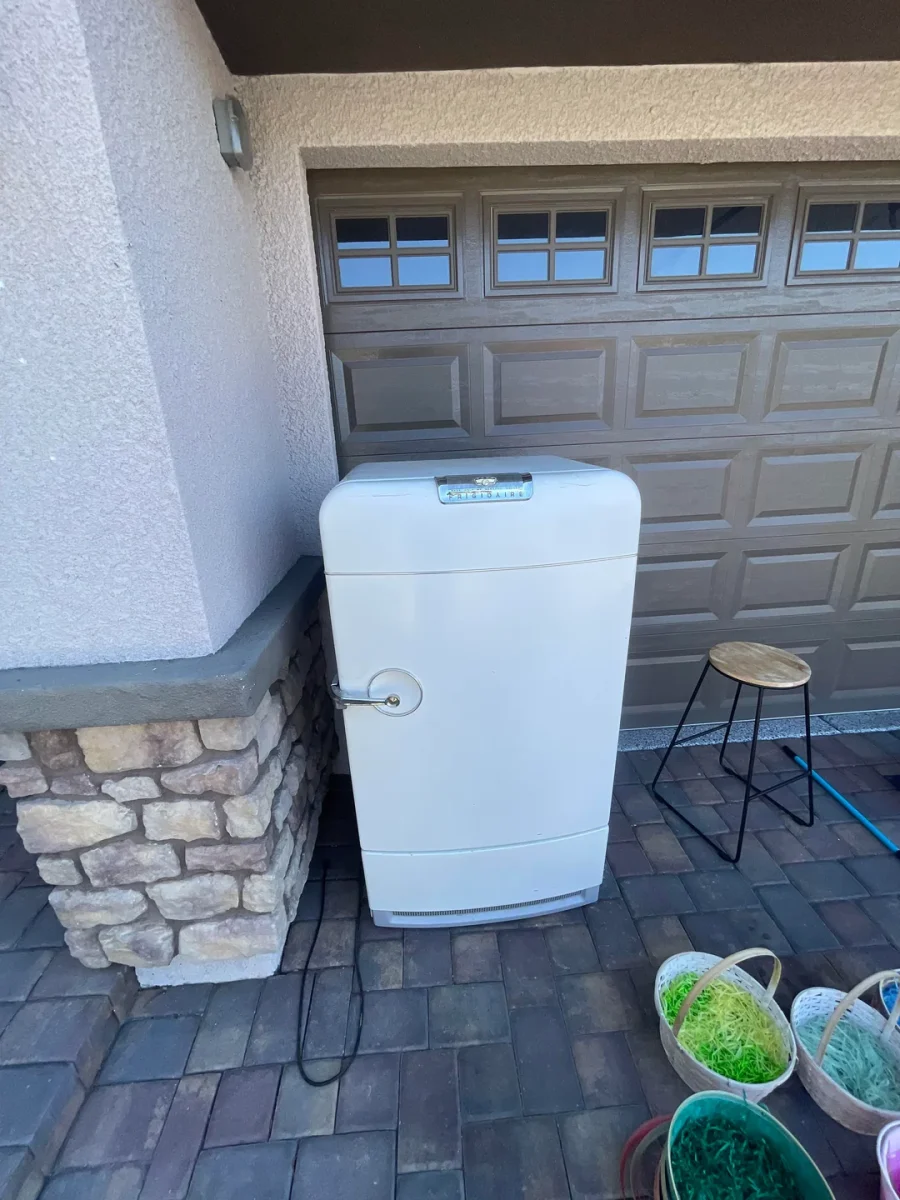 Starodávna chladnička pri garážových vrátach, obklopená kvetináčmi a stoličkou na chodníku.