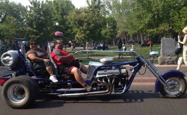 Motorka s unikátnym dizajnom a dvomi pasažiermi prechádza mestskou ulicou, obklopená zeleňou a okoloidúcimi.