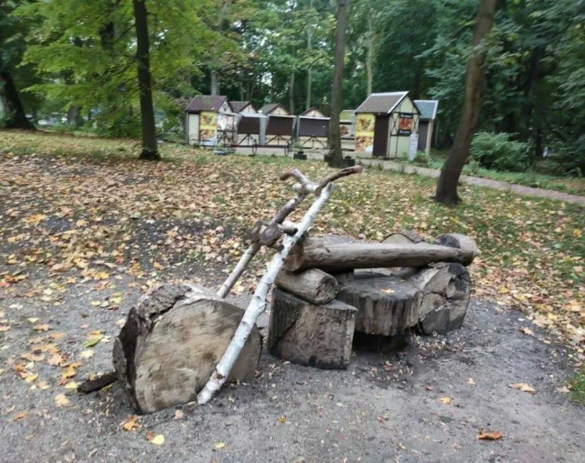 Drevená socha motorky v prírodnom prostredí parku, obklopená padajúcim lístím a stánkami v pozadí.