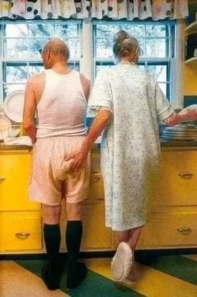 Starší pár umýva riad v kuchyni, žena láskyplne objíma muža v slnečnom svetle.