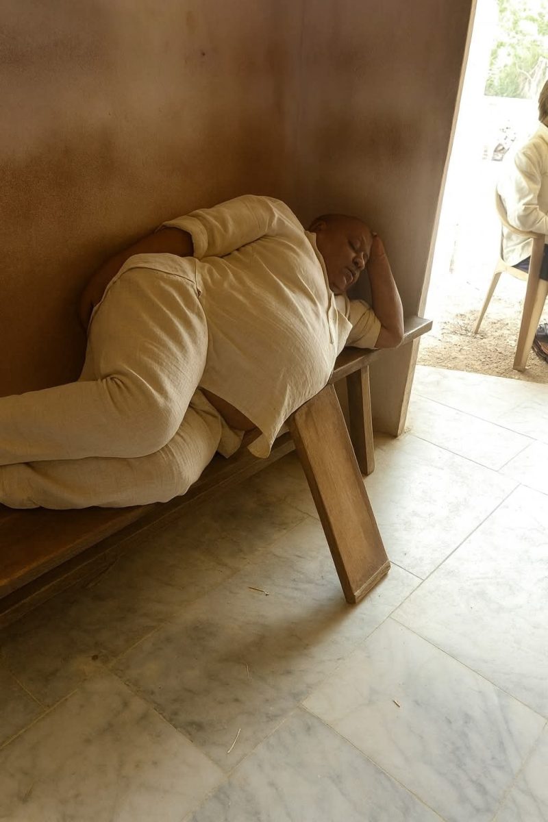 Osoba spí na lavičke v miestnosti, opretá o stenu, oblečená v bielom oblečení.