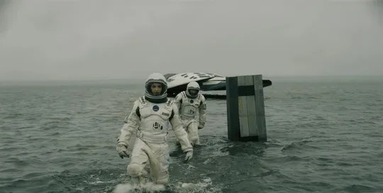 Astronauti kráčajú po vodnej planéte vedľa vesmírnej lode v hmlistskom prostredí.