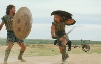 Dvaja bojovníci s mečmi a štítmi v akčnej scéne na piesočnom poli, v diaľke vidno voz.