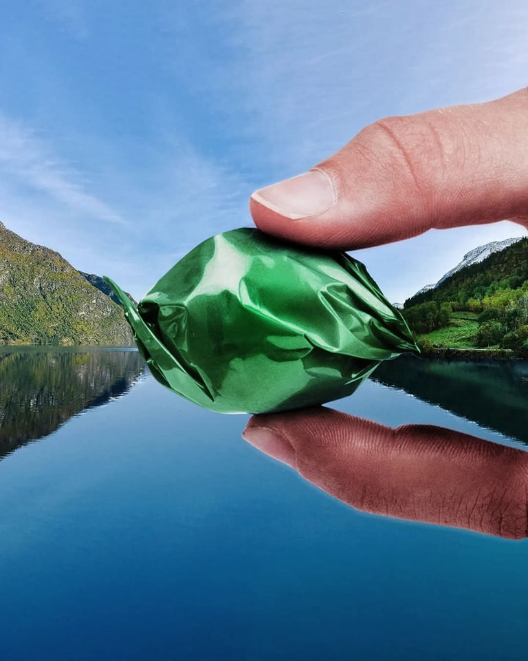 Prst držiaci zelený cukrík pred jazernou scenériou s odrazom hôr na vodnej ploche.