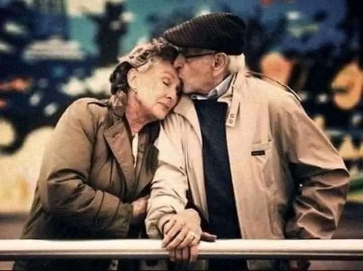 Starší pár v objatí na verejnom mieste, muž bozkáva ženu na čelo, symbol lásky a vernosti.