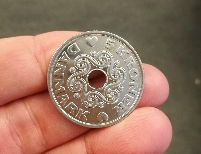 Dánska minca 5 korún s otvorom uprostred držiaca medzi prstami na sivej rozmazanej pozadí.