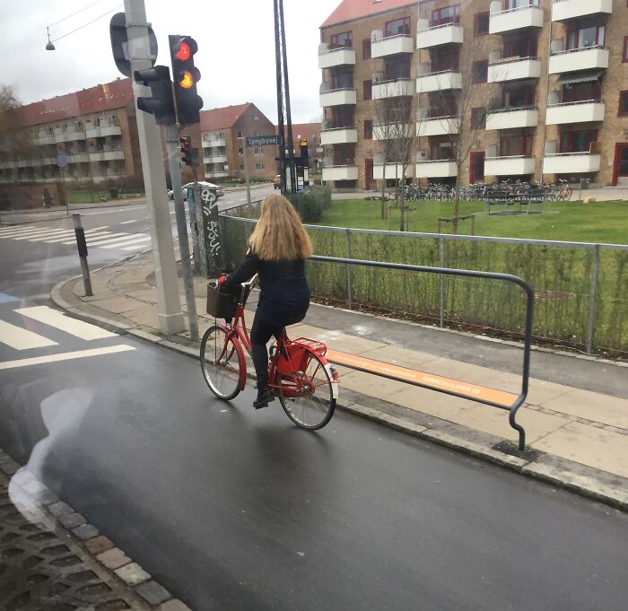 Žena na červenom bicykli na cyklochodníku v meste s modernými bytmi v pozadí.