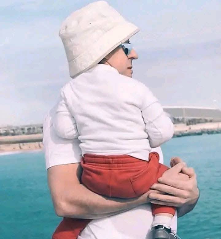 Osoba pri oceáne drží dieťa v červenom oblečení, slnečný deň na pláži.