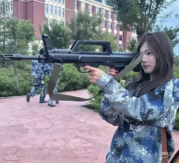 Mladá žena v uniformě s maketou zbrane v ruke, pozadie s budovou a stromami.