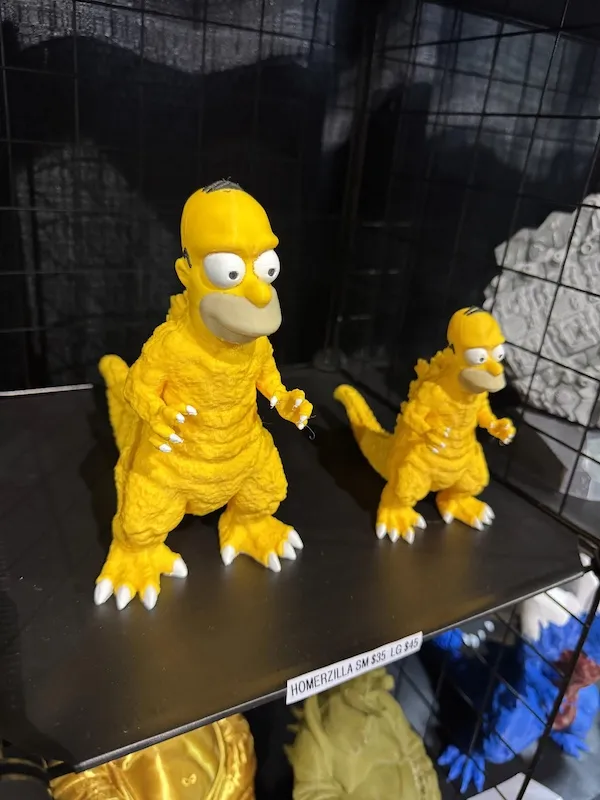 Dve plastové figúrky Homera v obleku Godzilly vystavené na polici.